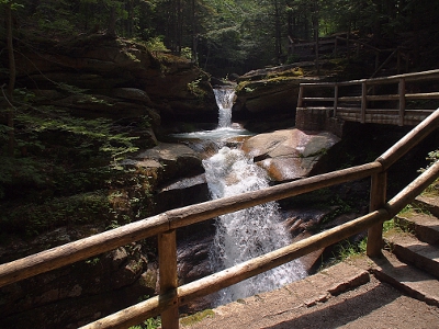 [A two tiered waterfall as seen through a post rail fence. The fence continues along the right of the image up to the ground next to the upper level of the falls.]